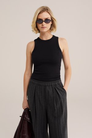 Blond naine kannab musta varrukateta toppi, tumeda triibulise laiade pükstega, mustad päikeseprillid, hoiab ühes käes tumedat pruuni jopet.