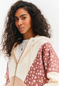 Bluza z kapturem wykonana z mieszaniny tkanin w kolorach kremowym, beżowym i różowym. Posiada akcenty z wzorem w cętki, sznurowadło oraz zamek błyskawiczny.