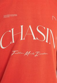 Raudona medvilninė marškinėliai su žodžiu "CHASING" ryškiomis baltomis raidėmis ir mažesniu šūkiu "Tradition Meets Innovation" išrašytu kursyvu apačioje.