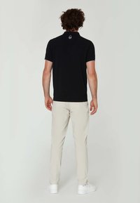 Camisa polo negra de manga corta, con cuello en contraste y logo en la parte trasera, combinada con pantalones de color claro y zapatillas blancas.