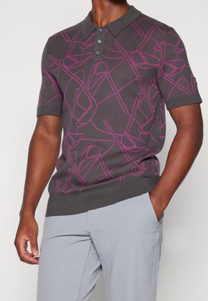 Mann trägt ein dunkelgraues kurzärmeliges Poloshirt mit pinken abstrakten Linienmustern und hellgraue Hose, eine Hand in der Tasche.