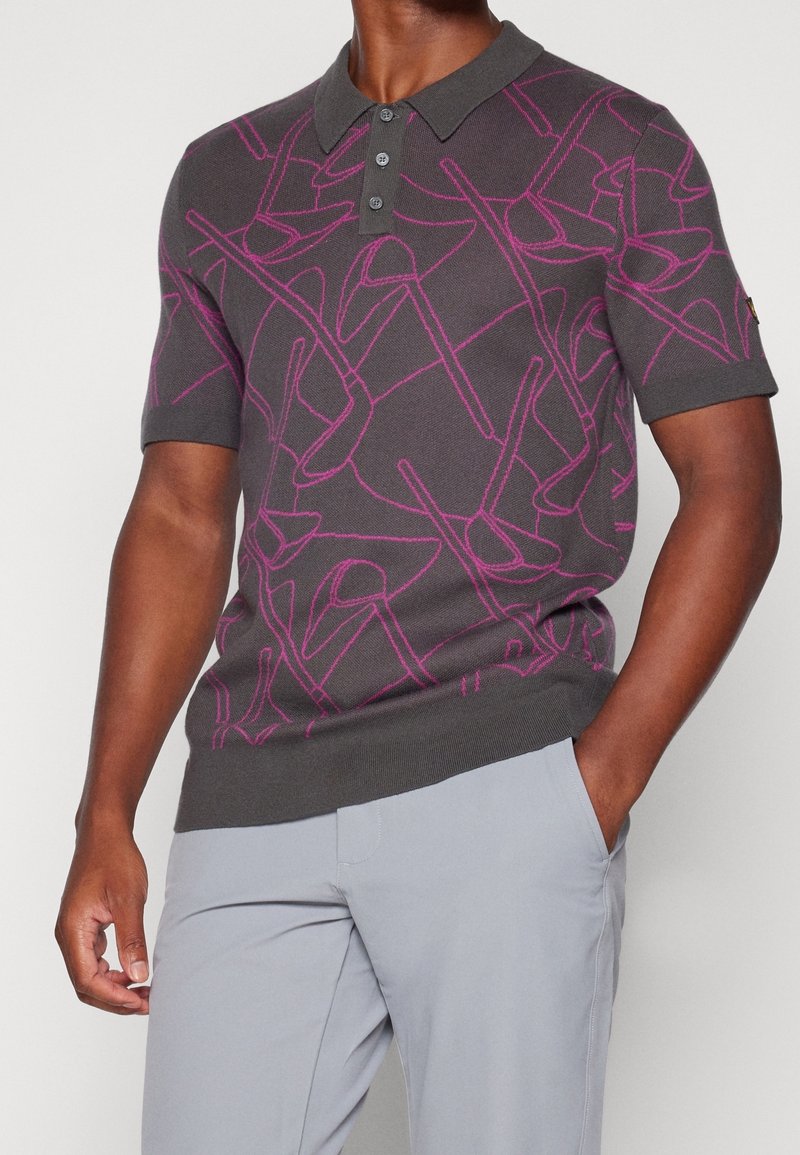 Homme portant un polo gris foncé à manches courtes avec des motifs abstraits de lignes roses et un pantalon gris clair, une main dans la poche.