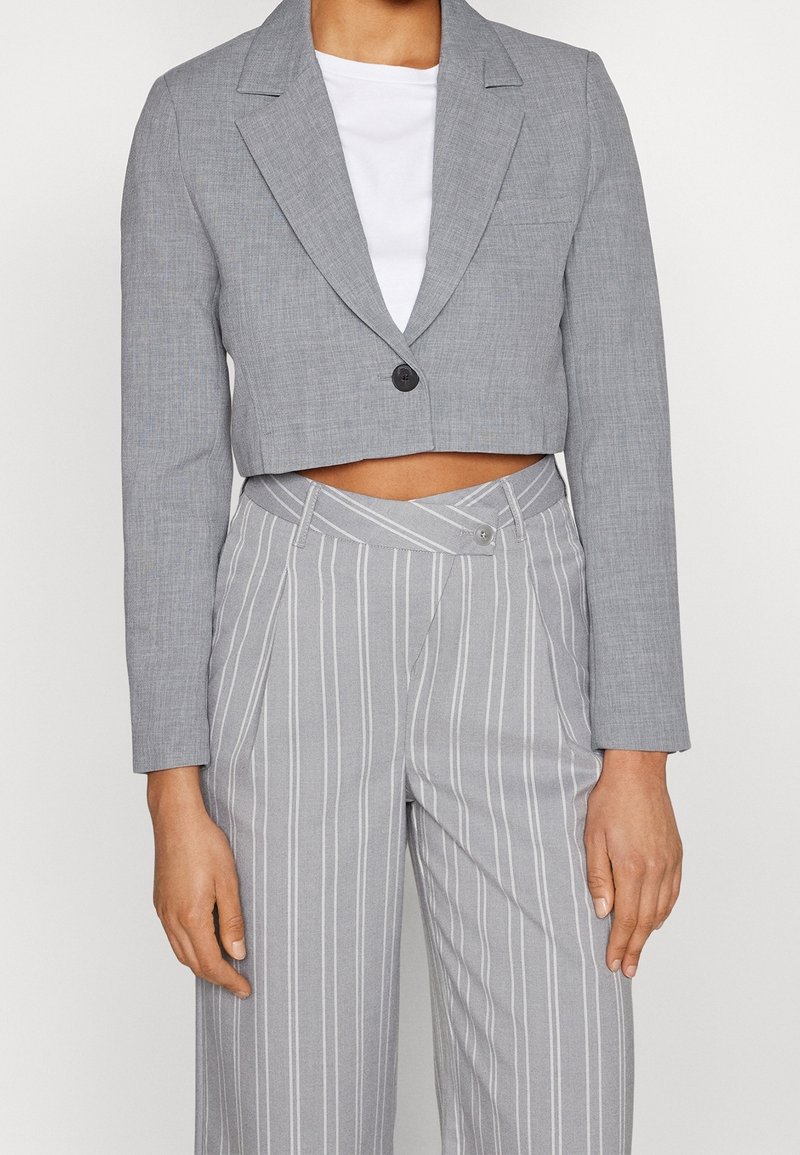 Femme portant un blazer gris court à un bouton sur un haut blanc, associé à un pantalon gris à fines rayures taille haute avec une taille boutonnée.