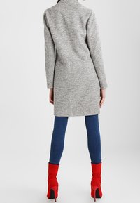 Langer grauer Mantel aus strukturiertem Stoff, mit einem Kragen und langen Ärmeln, kombiniert mit blauen Jeans und leuchtend roten Ankle-Boots.