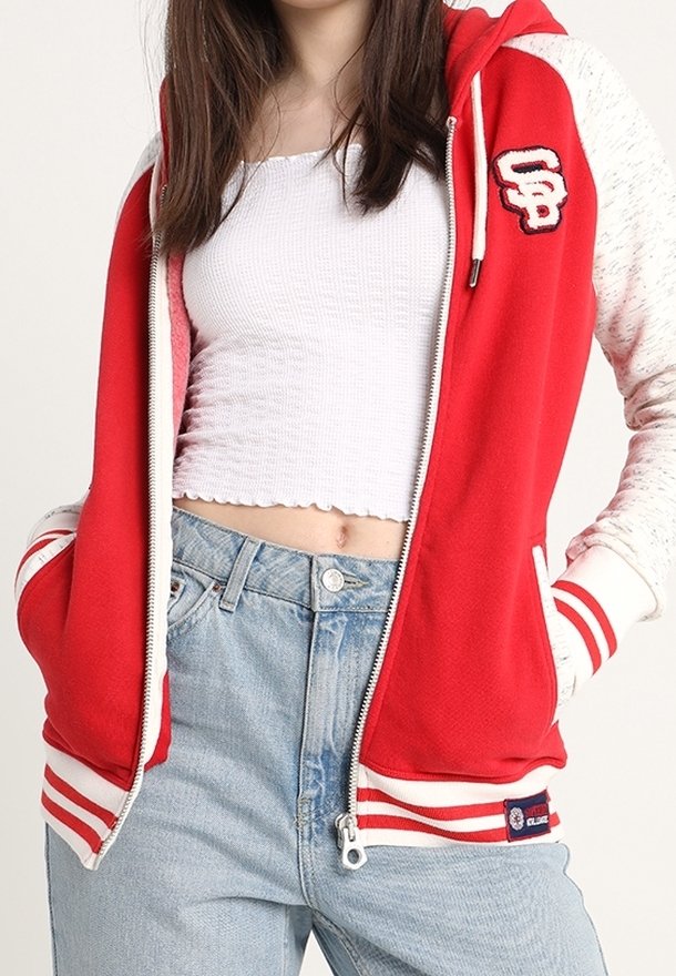 Sweat à capuche rouge avec des accents blancs, des poignets élastiques et un ourlet rayé. Présente un logo sur la poitrine et une fermeture éclair sur le devant. Porté par-dessus un crop top.