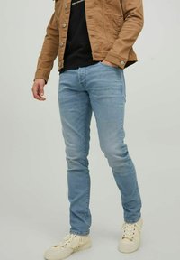 Los jeans azul claro tienen un corte ajustado y un diseño de cinco bolsillos. El modelo los combina con una chaqueta marrón y zapatillas blancas.