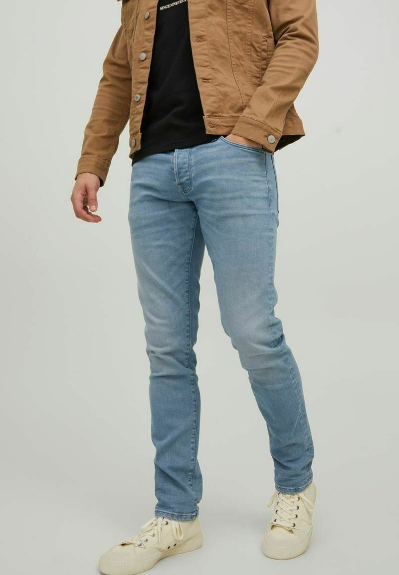 Los jeans azul claro tienen un corte ajustado y un diseño de cinco bolsillos. El modelo los combina con una chaqueta marrón y zapatillas blancas.