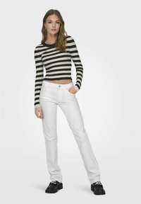 Maglione cropped a strisce nere e beige abbinato a jeans bianchi a gamba dritta. Il modello indossa scarpe con zeppa nere con lacci decorativi.