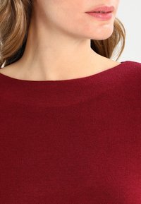 Burgundské pletené tričko s širokým výstřihem, hladkou texturou a přiléhavým střihem. Minimalistický design bez viditelného hardwaru nebo vzorů.