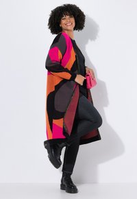 Femme souriante, portant un long cardigan coloré avec des motifs rose, orange et noir, un pantalon noir, des bottes noires et tenant une pochette rose.