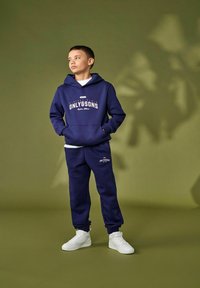 Marineblauwe hoodie met "ONLY&SONS" logo, gecombineerd met marineblauwe trainingsbroek. Model draagt witte sneakers. Soepele stof, casual pasvorm, geen patronen.