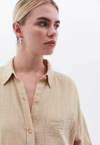 Camicia beige con bottoni, realizzata in un tessuto testurizzato, dotata di taschino sul petto e colletto classico, messa in risalto da piccoli bottoni di colore chiaro.