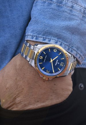 Orologio da polso con cassa argento e dorata, quadrante blu, numeri dorati e tre dettagli dorati. Indossato su un polso con tessuto di denim visibile.