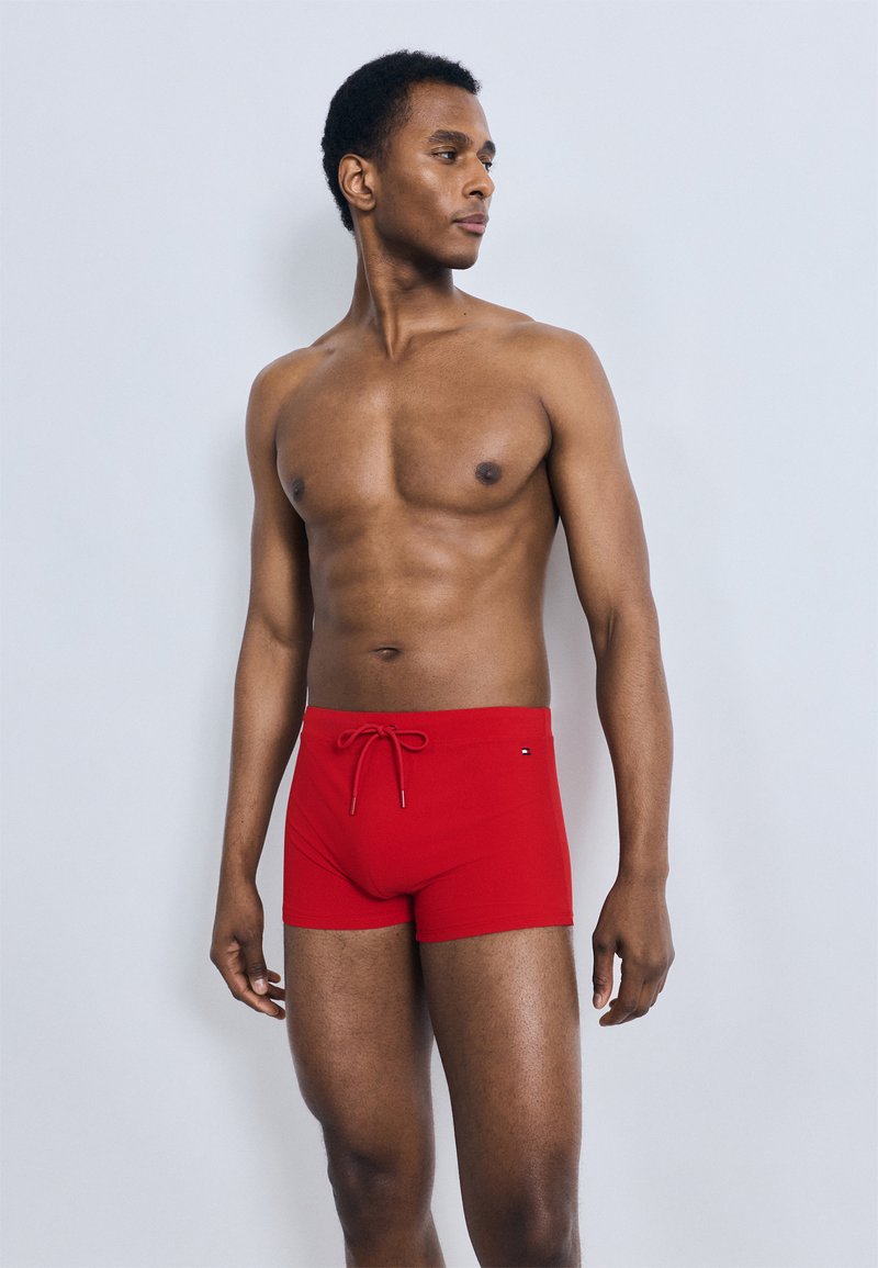 Κόκκινα μαγιό σορτς από λείο ύφασμα, με κορδόνι στη μέση, κοντό μήκος στο εσωτερικό, και λογότυπο στο πλάι.