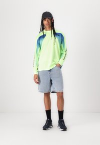 Neon grønn langermet skjorte med blå detaljer, denimshorts, svarte sokker og joggesko. Skjorten har en mesh tekstur og logo.