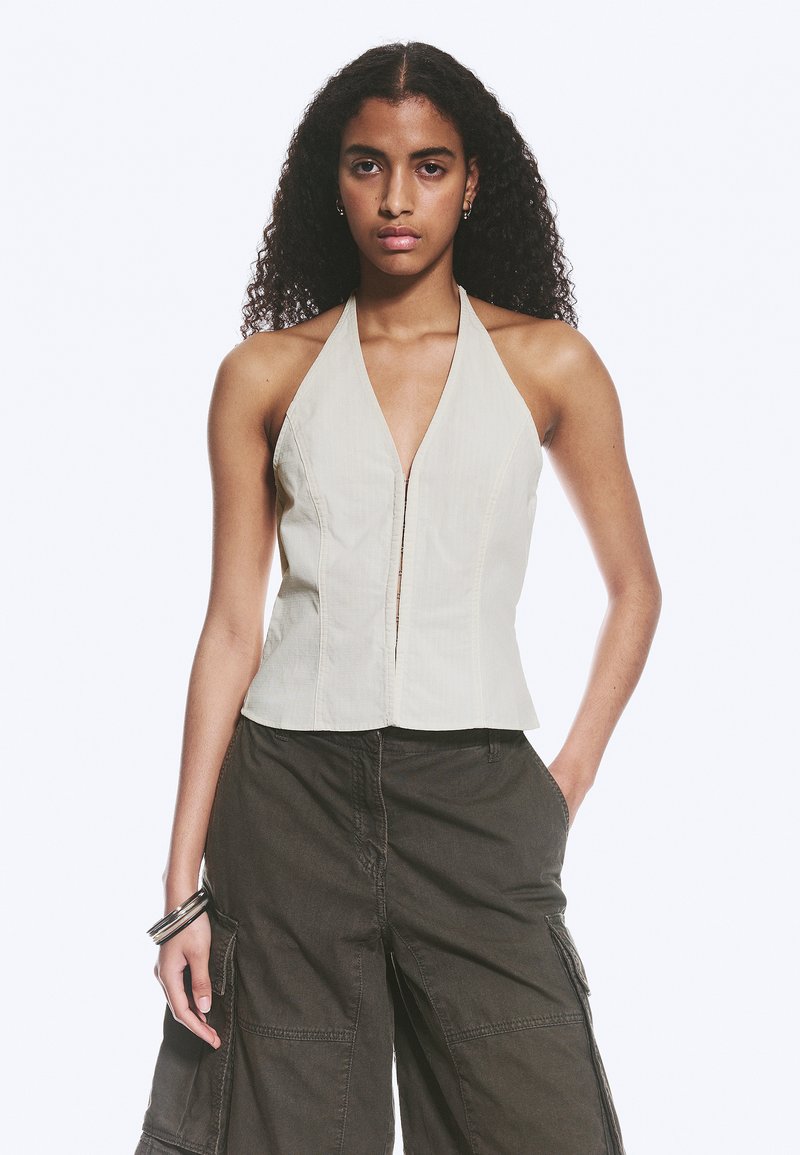 Jeune femme aux cheveux bouclés portant un haut beige à dos nu et un pantalon cargo foncé, posant avec une main dans la poche devant un fond blanc.