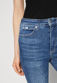 Blue denim jeans with a fitted design, featuring five pockets, a front button closure, and a small brand label on the waistband.