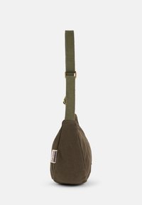Olive green canvas shoulder bag with a textured finish, tapered shape, adjustable strap, and a prominent fabric tag on the side.