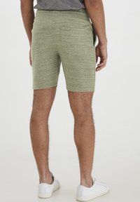 Shorts verde mélange con vita elastica, due tasche laterali e una texture liscia, abbinati a scarpe sportive bianche.