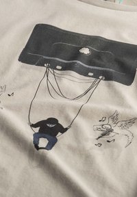 Beige katoenen t-shirt met een grafisch ontwerp van een zwarte cassetteband en een zittende figuur op een schommel, met muzieknoten en een gevleugeld paardenontwerp.