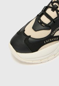 Musta ja beige lenkkari, jossa on verkkomateriaali yläosa, nahkaosuudet ja paksu, teksturoitu pohja. Huomattava logo-detaili sivussa.