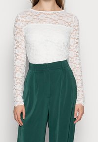 Haut à manches longues en dentelle blanche avec motif floral et corsage blanc uni ; associé à un pantalon taille haute vert, avec une texture lisse.