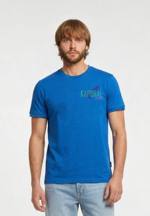 Man met lichtbruin haar en baard, die een blauw Kaporal t-shirt en lichtblauwe spijkerbroek draagt, staand tegen een effen achtergrond.