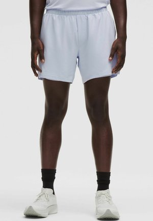 Personne portant un short de sport bleu clair, des chaussettes de sport noires et des chaussures de course blanches, se tenant devant un fond clair.