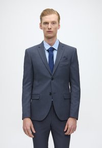 Fato azul marinho com um tecido texturizado, casaco de um só botão, lapelas com pico e calças combinando. Camisa azul clara e gravata escura.