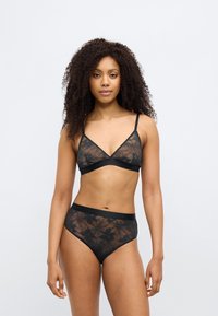 Γυναίκα με σγουρά μαλλιά που φοράει μαύρο διάφανο δαντελωτό σουτιέν με λουλούδια και σλιπ ψηλόμεσο, στέκεται μπροστά σε απλό ανοιχτό γκρι φόντο.