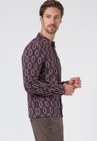 Camisa de manga larga y cierre de botones en color púrpura oscuro con un patrón geométrico en beige y rojo. Presenta un cuello puntiagudo y puños de botón.