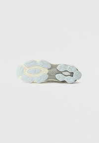 Sports shoe outsole features a light blue rubber grip with various textured patterns; the midsole is cream-colored, accentuating its shape.