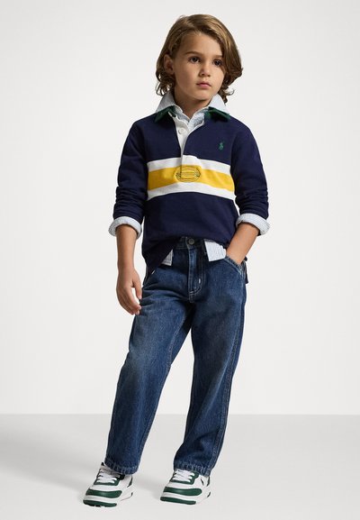 Polo de mangas longas em azul marinho com riscas amarelas e brancas, detalhes em verde, sobrepôsto a uma camisa de gola azul clara, acompanhado de jeans azuis e sapatilhas verdes.