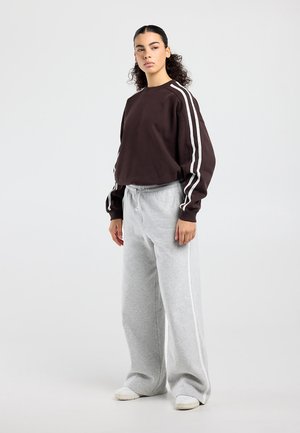 Junge Frau mit lockigem Haar, die einen dunkelbraunen Pullover mit weißen Streifen und lockere hellgraue Jogginghosen trägt, steht vor einem einfarbigen Hintergrund.