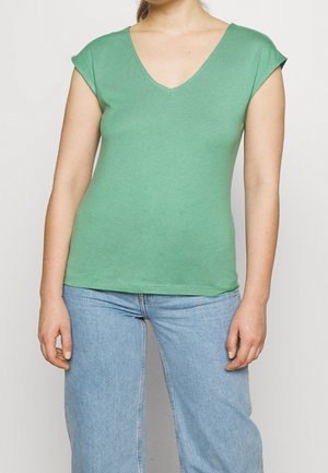 Femme portant un haut vert à manches courtes col V et un jean taille haute bleu clair, debout devant un fond uni.