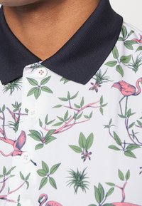 Polo à manches courtes avec un col bleu marine, présentant une base blanche et un imprimé botanique coloré avec des flamants roses. Tissu texturé.