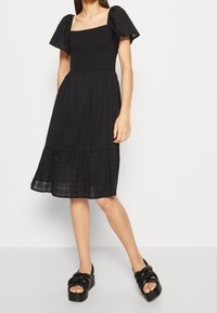 Schwarzes Kleid mit quadratischem Ausschnitt, Puffärmeln und gerafftem Oberteil. A-Linien-Rock mit mehrlagigem Saum. Kombiniert mit schwarzen Plateausandalen.