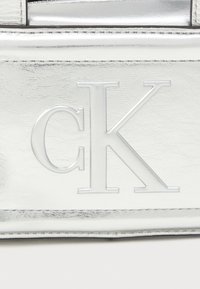 Srebrna torba z sztucznej skóry z wytłoczonym logo "CK". Gładka tekstura, prostokątny kształt oraz kontrastowe detale szwów.