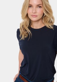 Tengerészkék, rövid ujjú póló puha anyagból, kerek nyakkivágással. A póló laza szabású, valamint farmernadrággal és barna övvel viselik.