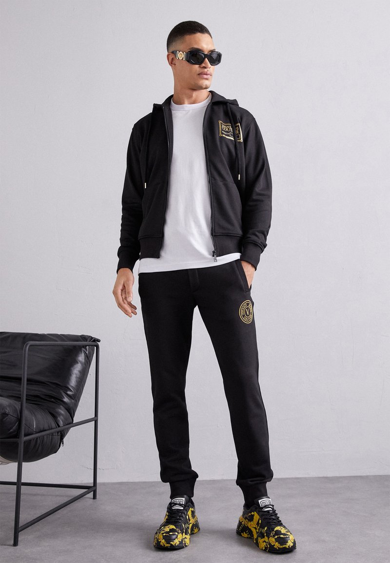 Zwarte zip-up hoodie met gouden logo, witte T-shirt, zwarte sweatpants met logo en voetbedekking met gele accenten. Zachte stof, ontspannen pasvorm.