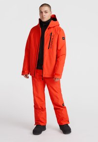 Veste de neige orange vif avec capuche, poches zippées et accents noirs, associée à un pantalon de neige orange assorti et des bottes noires.