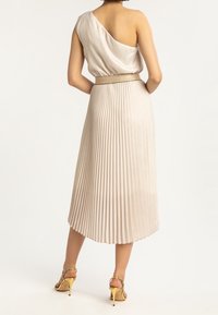 Jednoramienna plisowana sukienka z jasnobeżowego satynu o teksturowanym wykończeniu, z błyszczącym paskiem w talii i sandałami na złotym obcasie.