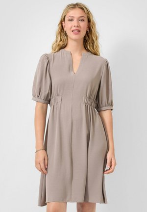 Junge Frau mit welligem blondem Haar, die ein knieumspielendes Kleid in Taupe mit Puffärmeln und V-förmigem Ausschnitt trägt.