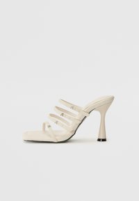 Sandales à talons hauts crème avec quatre fines lanières, bout carré, matériau texturé et un talon évasé avec un accent noir.