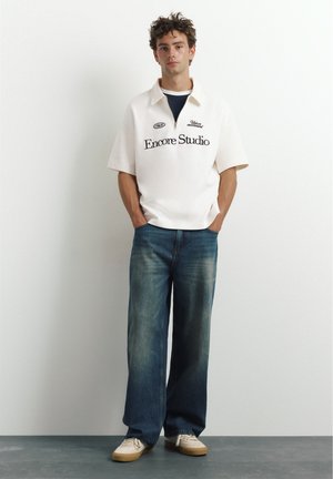 Jeune homme aux cheveux bouclés debout à l'intérieur portant une chemise blanche à manches courtes avec un texte, un jean bleu à jambes larges et des baskets blanches.