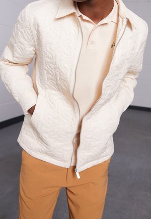 Quilted cream jacket with a zip closure, featuring an embossed pattern, paired with a light beige polo and tan pants.