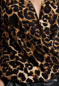 Blouse à imprimé léopard en nuances de noir et marron, présentant un design drapé et un tissu doux et texturé. Accents en cuir noir en bas.