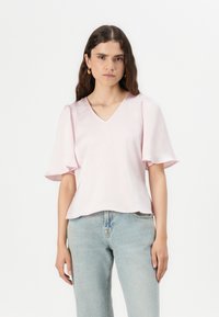 Top de color rosa claro con escote en V y mangas cortas abullonadas; hecho de una tela suave, con un corte holgado y un brillo sutil. Combinado con jeans de mezclilla claros.