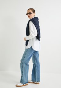 Jersey navy colocado sobre una camisa blanca, combinado con unos vaqueros de pierna ancha en un azul claro desgastado y zapatillas blancas con suelas marrones.