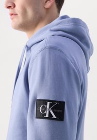Hanorac albastru deschis, fabricat dintr-un material moale, având o patch neagru cu text alb "Calvin Klein Jeans" pe mâneca stângă. Design simplu și cusături.